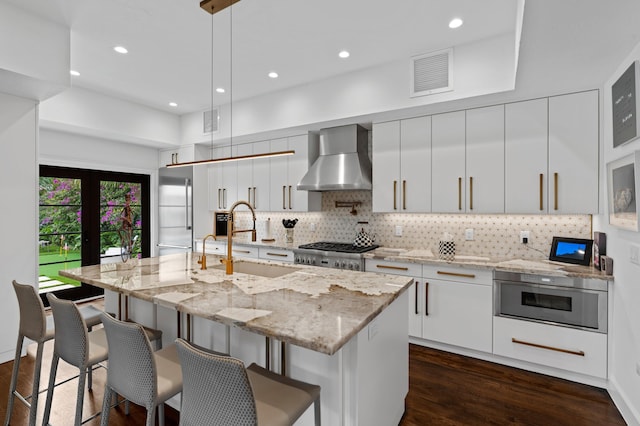 kitchen featuring wall chimney range hood, decorative light fixtures, an island with sink, and white cabinets