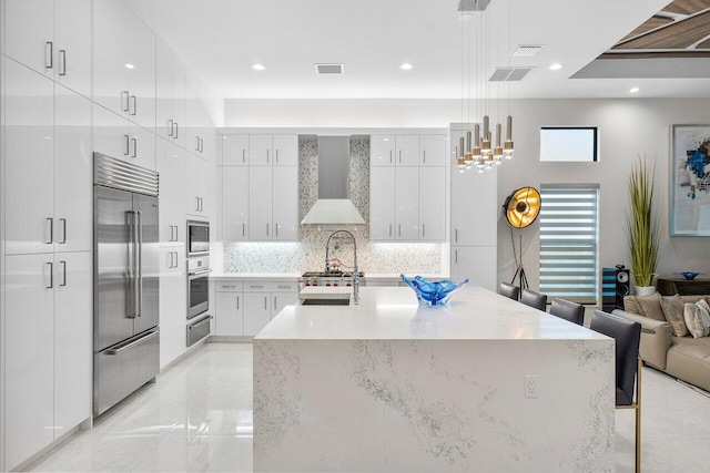 kitchen featuring built in appliances, wall chimney exhaust hood, white cabinetry, and decorative light fixtures