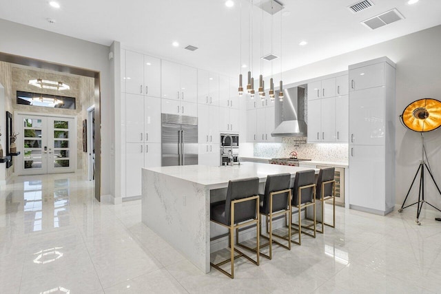 kitchen with pendant lighting, french doors, wall chimney range hood, an island with sink, and stainless steel built in refrigerator