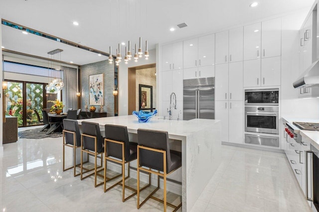 kitchen featuring a kitchen breakfast bar, pendant lighting, built in appliances, a large island with sink, and white cabinets