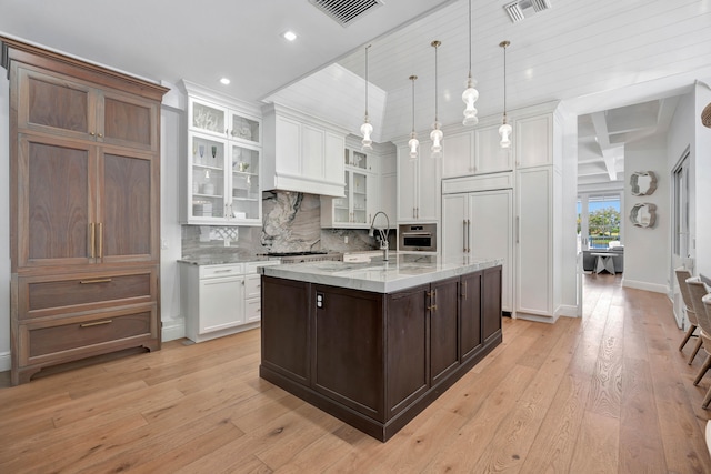 kitchen with light stone counters, white cabinetry, light wood-type flooring, decorative light fixtures, and an island with sink