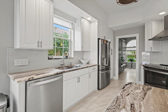 kitchen with light stone counters, stainless steel appliances, wall chimney range hood, sink, and white cabinets