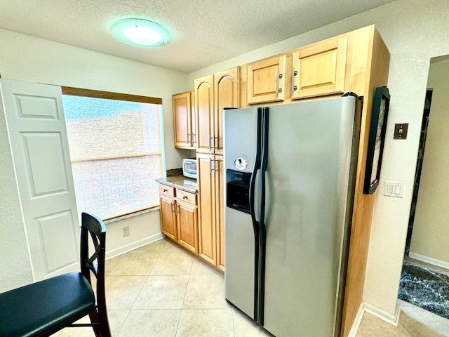 kitchen with light tile patterned floors, stainless steel fridge, a textured ceiling, dark stone counters, and light brown cabinets