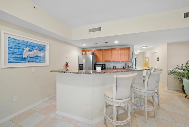 kitchen with stainless steel refrigerator with ice dispenser, dark stone counters, light tile patterned floors, and a breakfast bar area