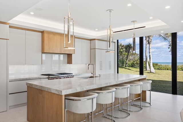 kitchen with white cabinetry, sink, hanging light fixtures, a raised ceiling, and a center island with sink