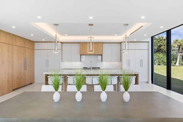 kitchen with pendant lighting, floor to ceiling windows, a tray ceiling, white cabinets, and a large island with sink