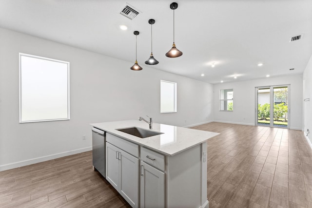 kitchen featuring a kitchen island with sink, sink, hanging light fixtures, stainless steel dishwasher, and light stone counters