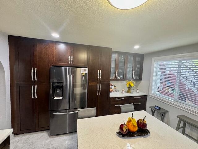 kitchen with light tile patterned flooring, stainless steel fridge with ice dispenser, dark brown cabinets, a textured ceiling, and a kitchen breakfast bar