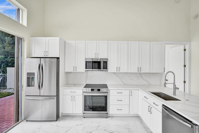 kitchen featuring a towering ceiling, light stone counters, stainless steel appliances, sink, and white cabinetry