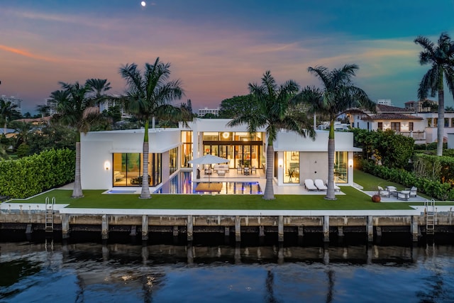 back house at dusk featuring a swimming pool, a lawn, a water view, and a patio area