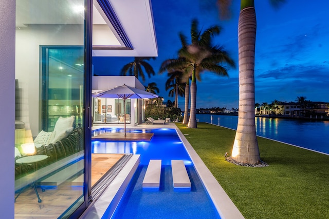 pool at dusk featuring a patio, a water view, and a lawn