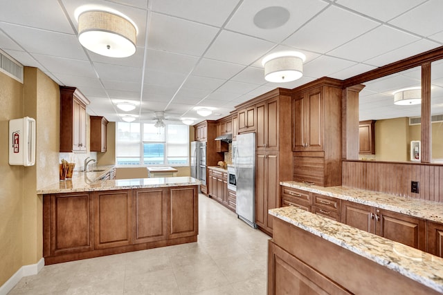 kitchen with stainless steel fridge, kitchen peninsula, light stone countertops, a drop ceiling, and ceiling fan