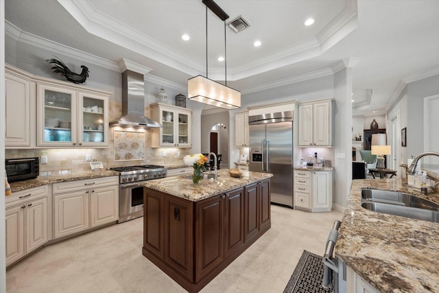 kitchen featuring high end appliances, glass insert cabinets, wall chimney range hood, pendant lighting, and a sink