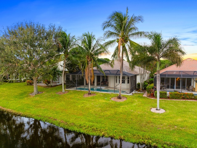 back house at dusk featuring a lawn, a lanai, and a water view