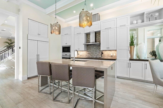 kitchen featuring built in appliances, a center island with sink, sink, hanging light fixtures, and wall chimney range hood