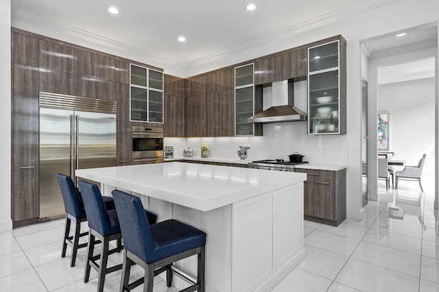 kitchen with appliances with stainless steel finishes, ornamental molding, wall chimney exhaust hood, a breakfast bar, and a kitchen island