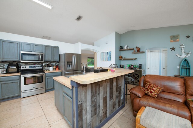 kitchen with a kitchen island with sink, vaulted ceiling, appliances with stainless steel finishes, tasteful backsplash, and light tile patterned flooring