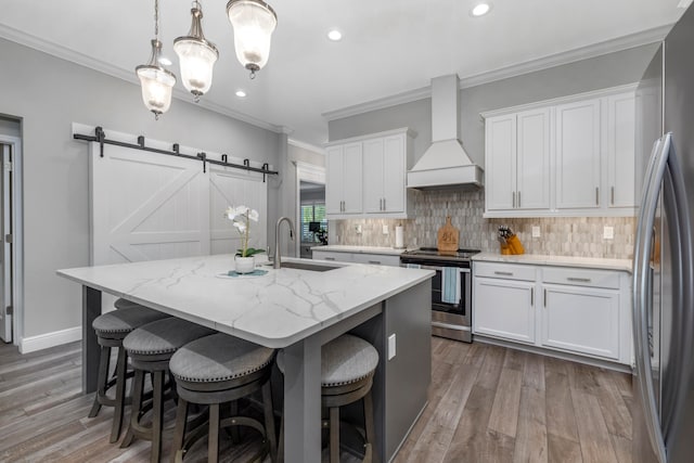 kitchen with custom range hood, stainless steel appliances, a barn door, white cabinetry, and an island with sink