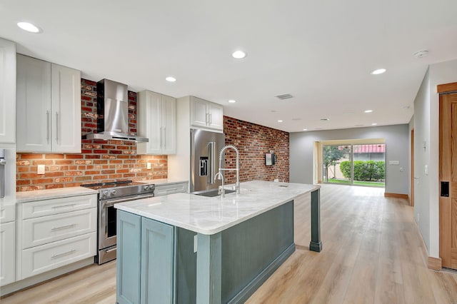 kitchen featuring stainless steel appliances, light stone countertops, wall chimney exhaust hood, an island with sink, and light hardwood / wood-style flooring