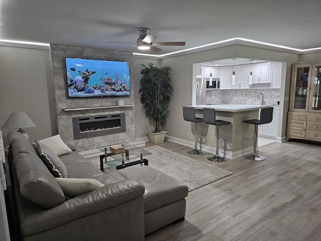 living room featuring ceiling fan, crown molding, sink, light hardwood / wood-style floors, and a stone fireplace