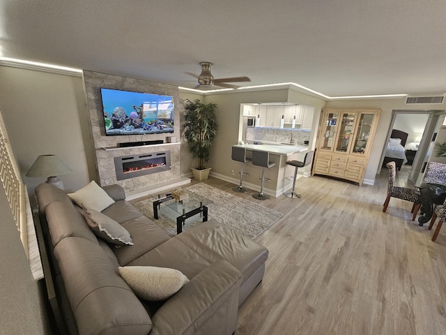 living room featuring visible vents, light wood-style floors, ceiling fan, a stone fireplace, and baseboards