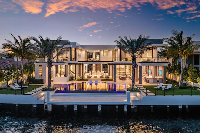 back house at dusk with a water view, a balcony, a patio, a lawn, and outdoor lounge area