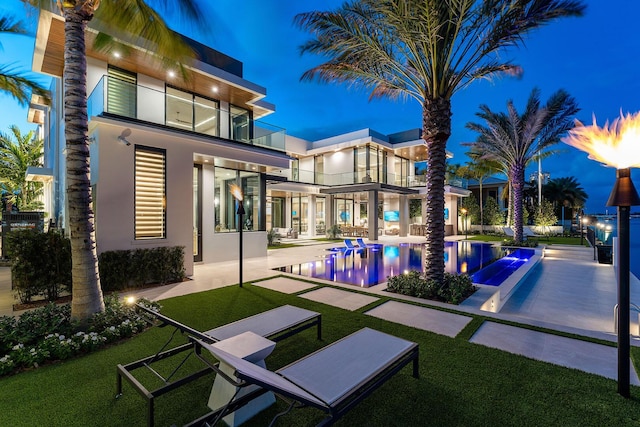1520 Royal Palm Way, Boca Raton, FL 33432 - back of house at night with a balcony, stucco siding, an infinity pool, a patio area, and a lawn