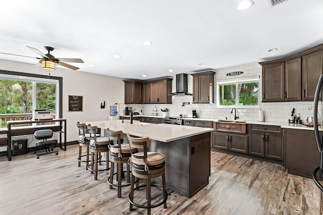 kitchen featuring a wealth of natural light, light hardwood / wood-style floors, wall chimney range hood, and an island with sink