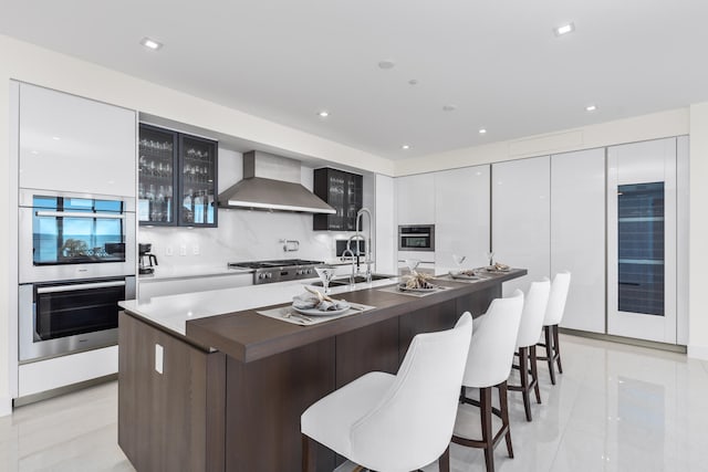 kitchen with stainless steel appliances, white cabinetry, tasteful backsplash, an island with sink, and wall chimney range hood