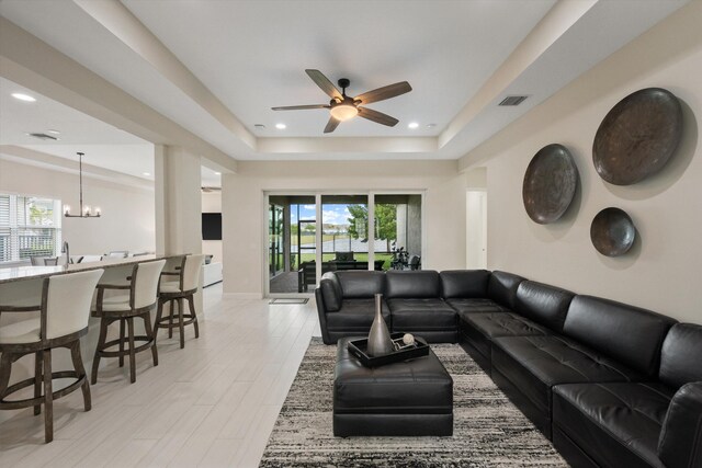 living room featuring sink, a tray ceiling, ceiling fan with notable chandelier, and light hardwood / wood-style flooring