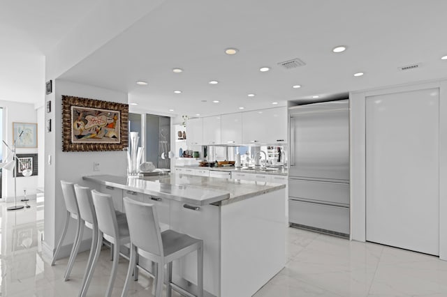 kitchen with built in refrigerator, sink, a kitchen breakfast bar, light stone countertops, and white cabinetry