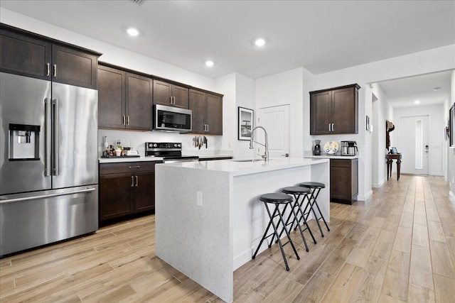 kitchen with a center island with sink, appliances with stainless steel finishes, dark brown cabinetry, sink, and light hardwood / wood-style flooring