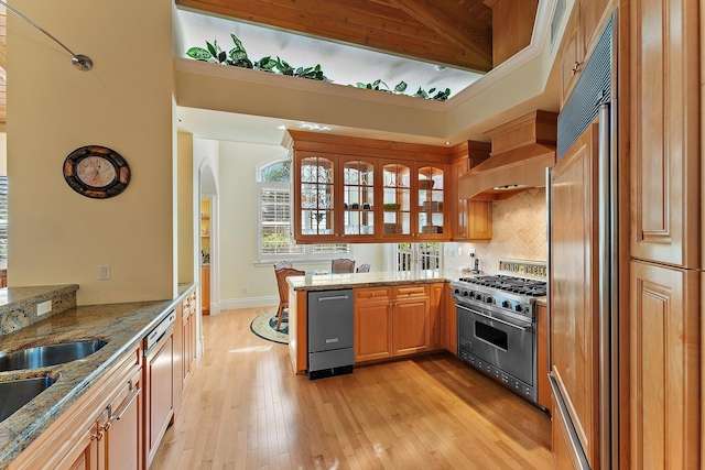 kitchen featuring high quality appliances, light stone counters, kitchen peninsula, wall chimney exhaust hood, and light wood-type flooring