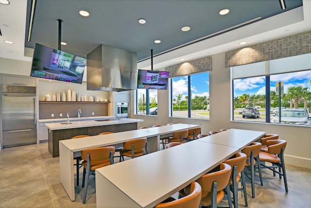 kitchen with wall chimney range hood, appliances with stainless steel finishes, a breakfast bar area, and a kitchen island with sink