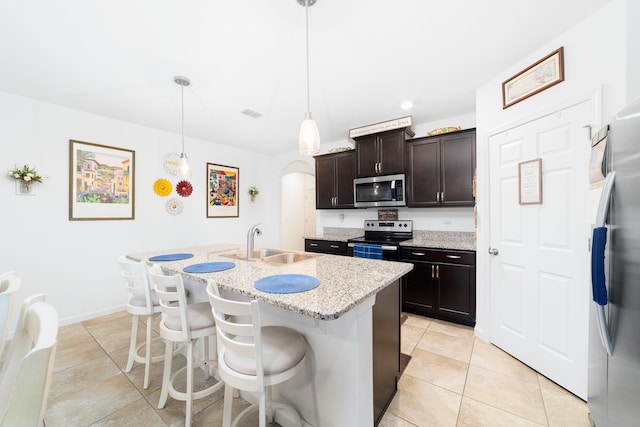 kitchen featuring hanging light fixtures, light stone countertops, an island with sink, appliances with stainless steel finishes, and dark brown cabinets