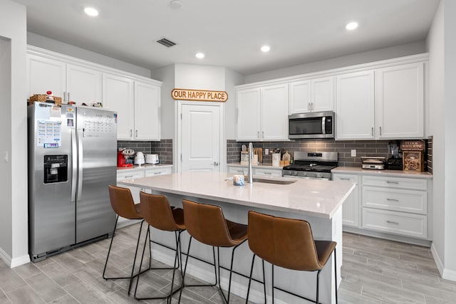 kitchen with light hardwood / wood-style floors, stainless steel appliances, a center island with sink, backsplash, and white cabinetry