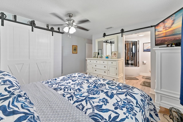 bedroom with ensuite bath, a textured ceiling, a barn door, light hardwood / wood-style floors, and ceiling fan