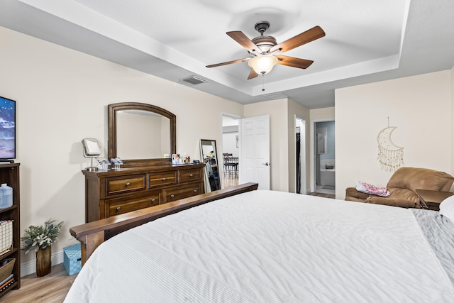 bedroom with ensuite bathroom, light wood-type flooring, ceiling fan, and a tray ceiling