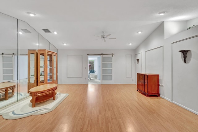 unfurnished living room featuring ceiling fan, a barn door, light hardwood / wood-style flooring, and vaulted ceiling