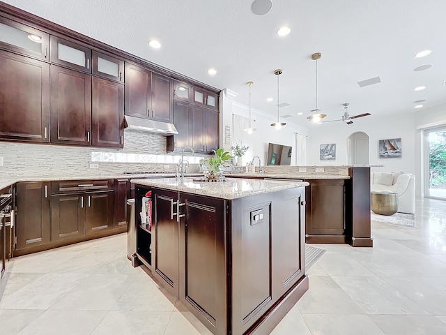 kitchen featuring light stone counters, dark brown cabinets, hanging light fixtures, and an island with sink