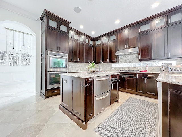 kitchen featuring light stone counters, ornamental molding, stainless steel appliances, backsplash, and dark brown cabinets