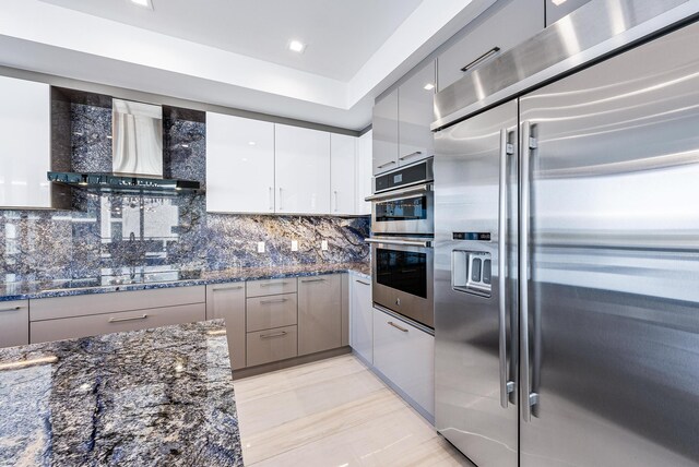 kitchen with stainless steel appliances, backsplash, wall chimney range hood, and dark stone countertops