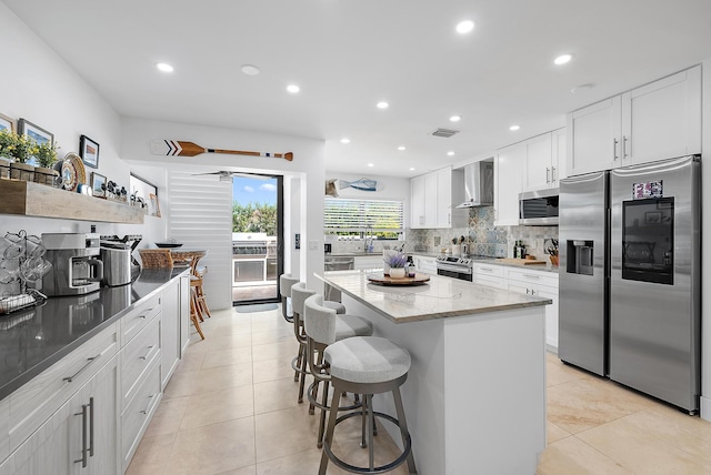 kitchen featuring a center island, wall chimney range hood, light stone countertops, white cabinetry, and stainless steel appliances