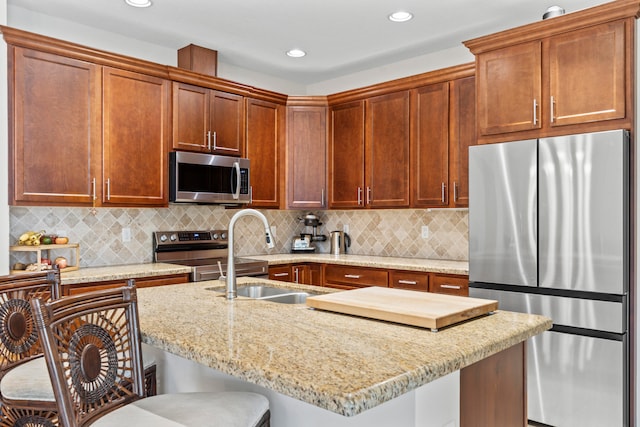 kitchen with stainless steel appliances, decorative backsplash, a kitchen island with sink, light stone counters, and a breakfast bar area