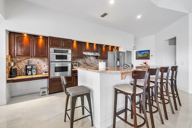 kitchen with appliances with stainless steel finishes, a kitchen breakfast bar, light stone countertops, light tile patterned floors, and backsplash