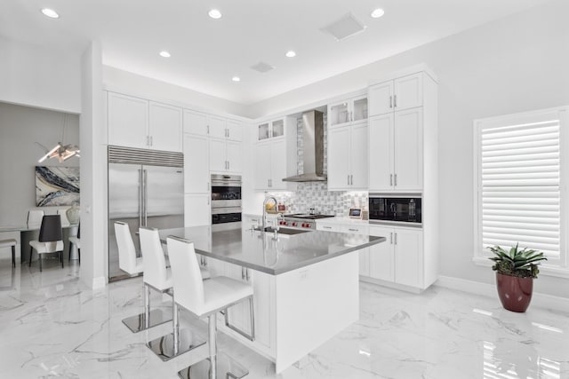 kitchen with an island with sink, sink, a breakfast bar area, built in appliances, and wall chimney range hood