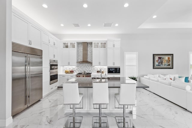 kitchen with wall chimney exhaust hood, a breakfast bar, white cabinetry, built in appliances, and a center island with sink