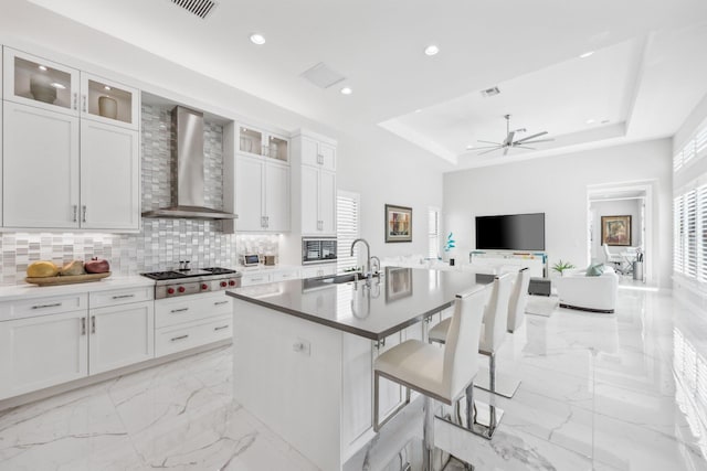 kitchen with a tray ceiling, a kitchen breakfast bar, wall chimney range hood, a kitchen island with sink, and white cabinets