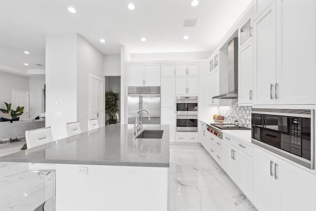 kitchen featuring wall chimney range hood, sink, a spacious island, built in appliances, and white cabinets