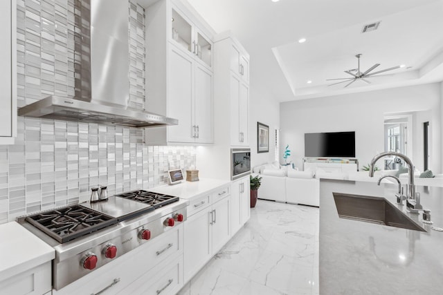 kitchen with sink, white cabinets, stainless steel gas cooktop, a tray ceiling, and wall chimney range hood